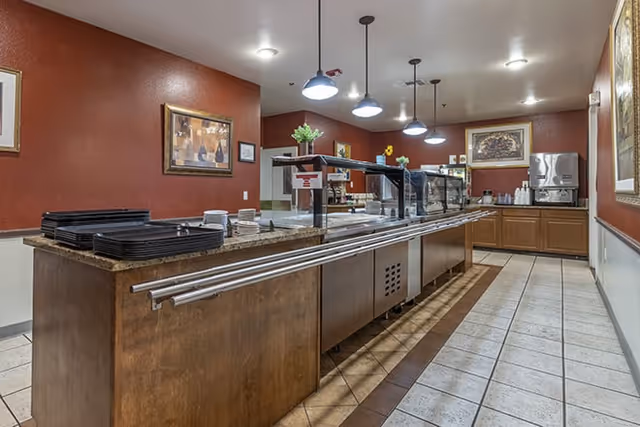 Interior view of a dining area with a long buffet counter featuring trays, plates, and a sneeze guard. The walls are painted red with framed artwork, and there are pendant lights hanging from the ceiling. A beverage dispenser is visible in the background.