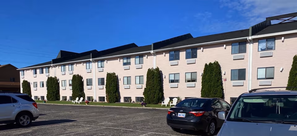 Three-story light-colored apartment building with trimmed evergreens, a parking lot with cars in front, and a clear blue sky.