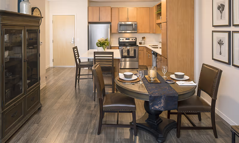 Open-concept kitchen and dining area with a round set table, island seating, stainless steel appliances, and wooden cabinetry.