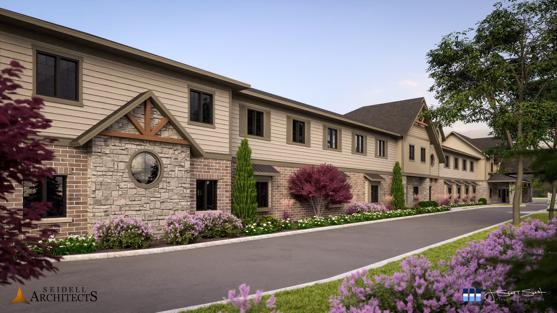Exterior view of a two-story senior living facility building with a combination of stone, brick, and siding on the facade. The building is surrounded by landscaped greenery including trees, shrubs, and flowering plants along a paved driveway or road.