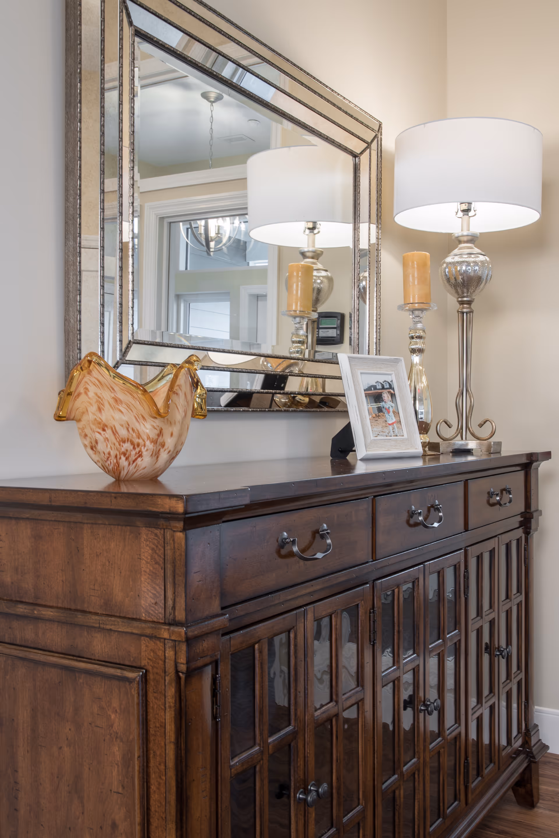 A wooden sideboard with glass-paneled cabinet doors and three drawers. On top of the sideboard are a decorative glass rooster, a framed photo, a silver candlestick with a yellow candle, and a silver table lamp with a white lampshade. Above the sideboard is a large, decorative mirror reflecting part of the room including a chandelier and windows.