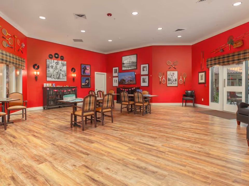 A spacious room with bright red walls and wood flooring, furnished with several tables and chairs arranged for group seating. The walls are decorated with vintage tennis rackets, black and white photographs, and vinyl records. Large windows with plaid valances allow natural light to enter, and there is a door leading outside. The room has a cozy and inviting atmosphere.