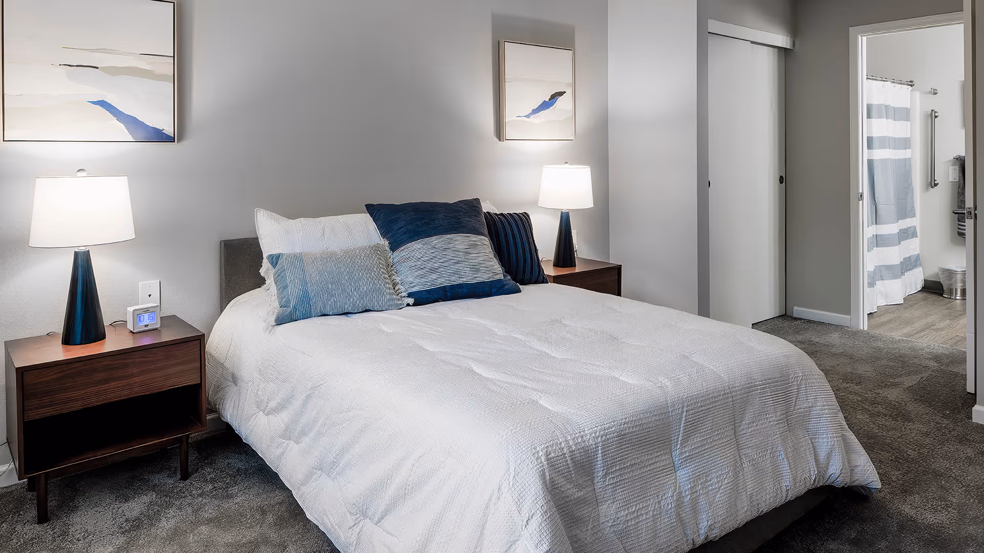 A neatly made bed with white bedding and multiple blue and gray pillows in a bedroom. There are two wooden nightstands on either side of the bed, each with a modern lamp. Abstract artwork hangs above each nightstand. In the background, there is an open doorway leading to a bathroom with a striped shower curtain and a closet with sliding doors.