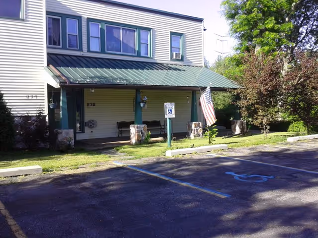 Exterior view of a two-story building with a green metal roof over a porch area. The porch has benches and stone pillars, an American flag is displayed, and there is a handicapped parking space in front. Trees and greenery surround the building.