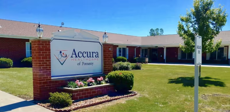 Exterior view of Accura HealthCare of Pomeroy building with a large sign in front surrounded by grass, bushes, and flowers under a clear blue sky.