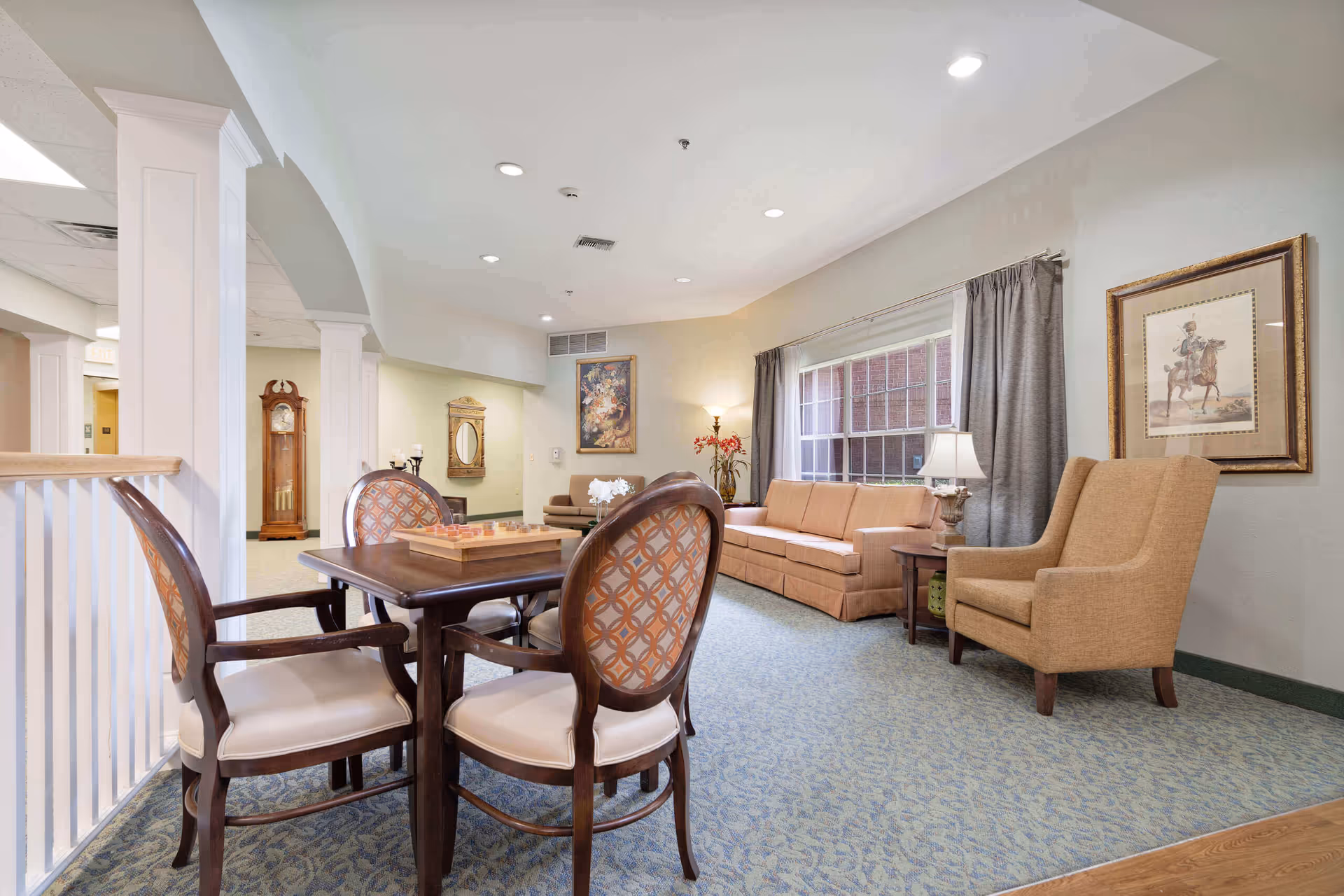 A bright and cozy living room area in a senior living facility featuring a wooden table with four upholstered chairs, a beige sofa, a matching armchair, a side table with a lamp, framed artwork on the walls, a large window with gray curtains, and a grandfather clock in the background.