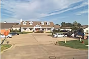 Front exterior of a two-story retirement community building with a circular driveway, parked cars, and landscaping under a partly cloudy sky.