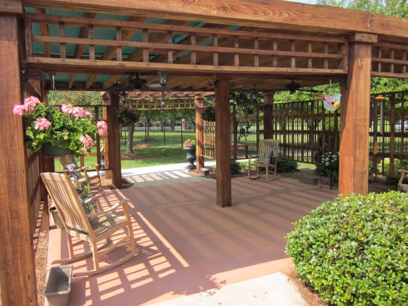 Wooden pergola-covered patio with rocking chairs, hanging flower baskets, and surrounding greenery.