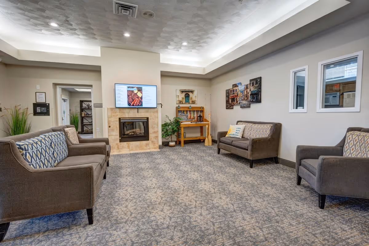 A cozy living room area in South Hill Village featuring three brown sofas with patterned cushions arranged around a carpeted floor. A fireplace with a mounted flat-screen TV above it is centered on the far wall. There are decorative plants, a wooden table with a painting above it, and two windows on the right wall showing another room.