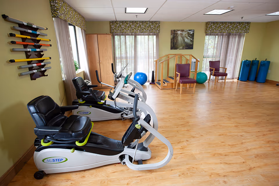 A bright exercise room with three NuStep recumbent exercise bikes lined up on a wooden floor. The room has light green walls, large windows with patterned valances and sheer curtains, two purple chairs, a small wooden staircase for physical therapy, blue exercise balls, and foam rollers in the corner. A nature-themed picture hangs on the wall.