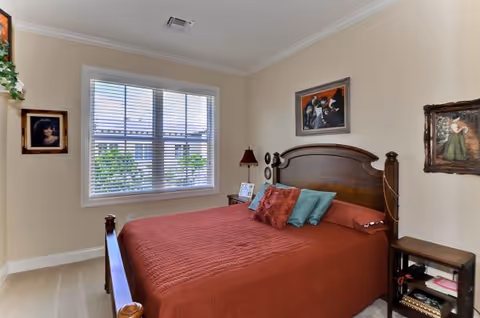A bedroom with a wooden bed frame and a red bedspread, decorated with multiple pillows. The room has beige walls with three framed paintings hanging on them. A window with white blinds lets in natural light, and there is a small wooden nightstand with a lamp and picture frame beside the bed.