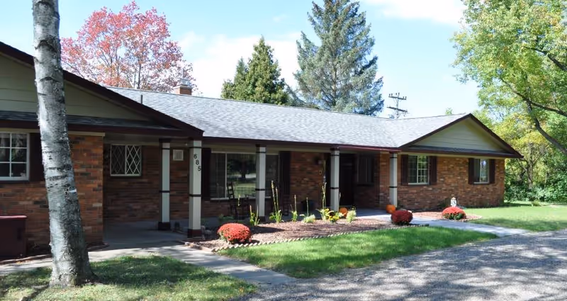 Exterior view of a single-story brick building with a covered porch, surrounded by green grass, trees, and a gravel driveway under a clear blue sky.