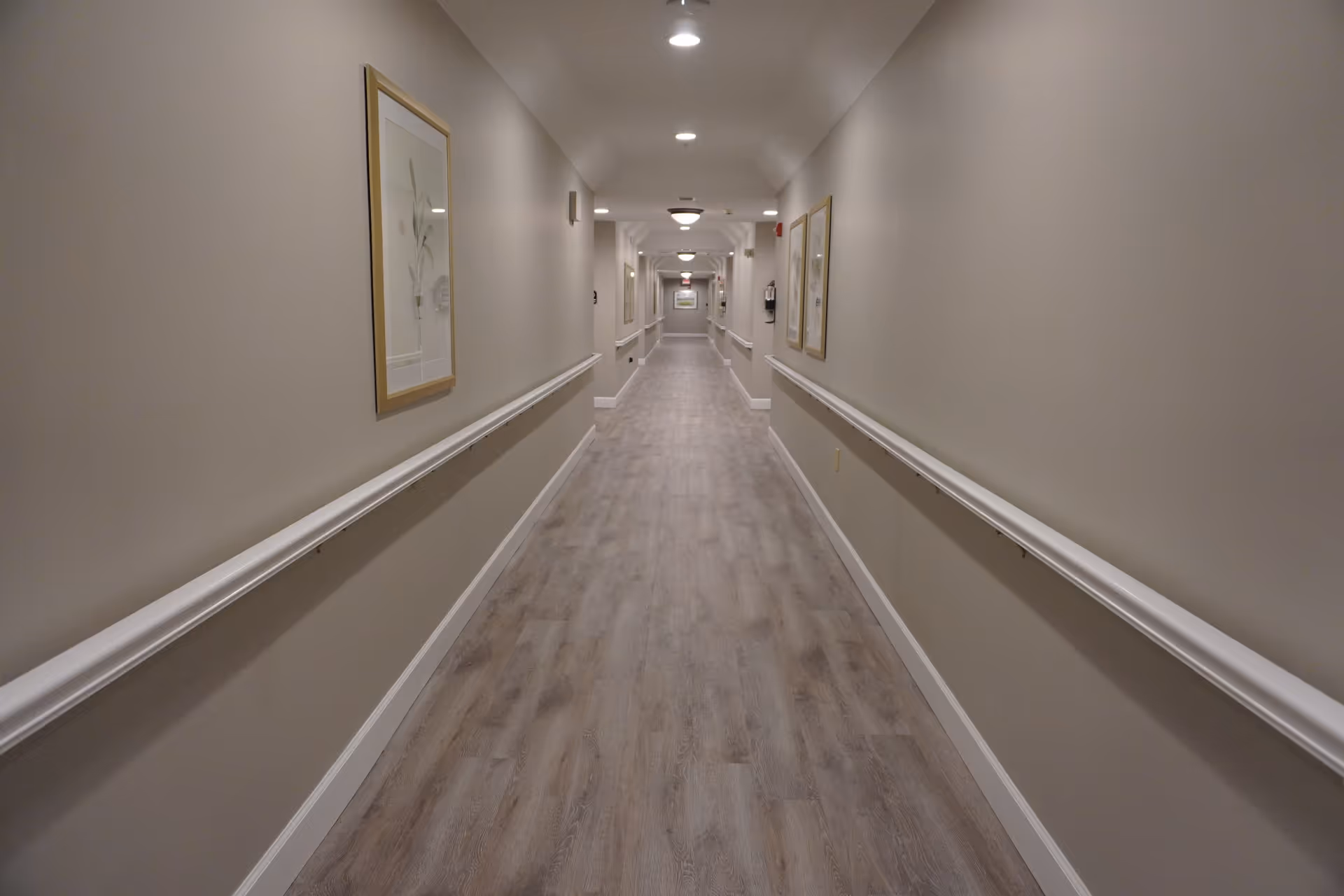 A long, well-lit hallway with light-colored walls and wood flooring. The hallway has handrails on both sides and framed artwork hanging on the walls. Ceiling lights are evenly spaced along the corridor.