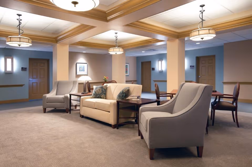 A spacious and well-lit common area in a senior living facility featuring beige and gray upholstered chairs and a beige sofa with decorative pillows arranged around wooden side tables. The room has beige walls with wooden trim, multiple closed doors, framed artwork, and ceiling light fixtures.