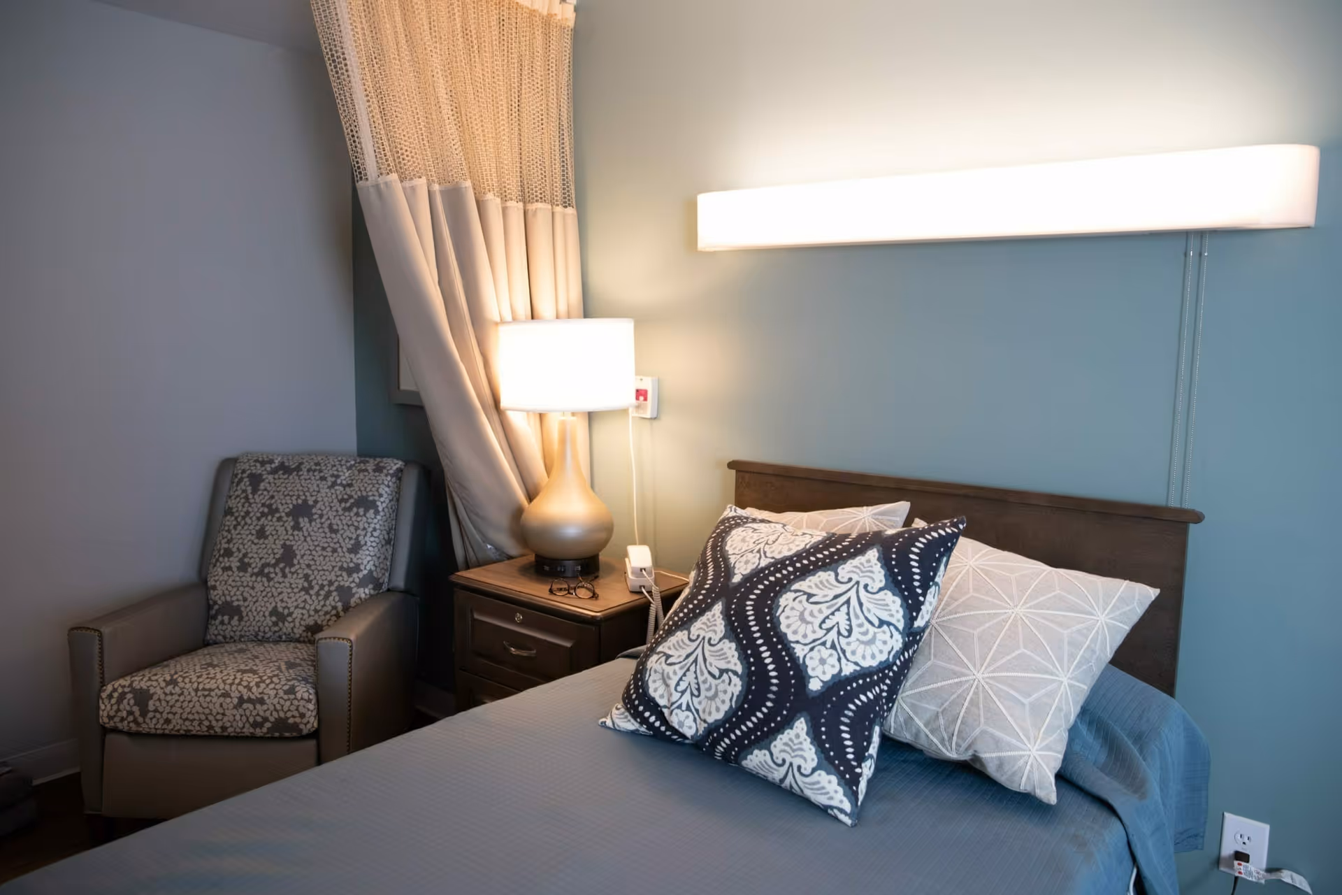 A cozy bedroom with a neatly made bed featuring three decorative pillows. Next to the bed is a wooden nightstand with a modern lamp emitting warm light. A patterned armchair is positioned in the corner near a window with beige curtains.