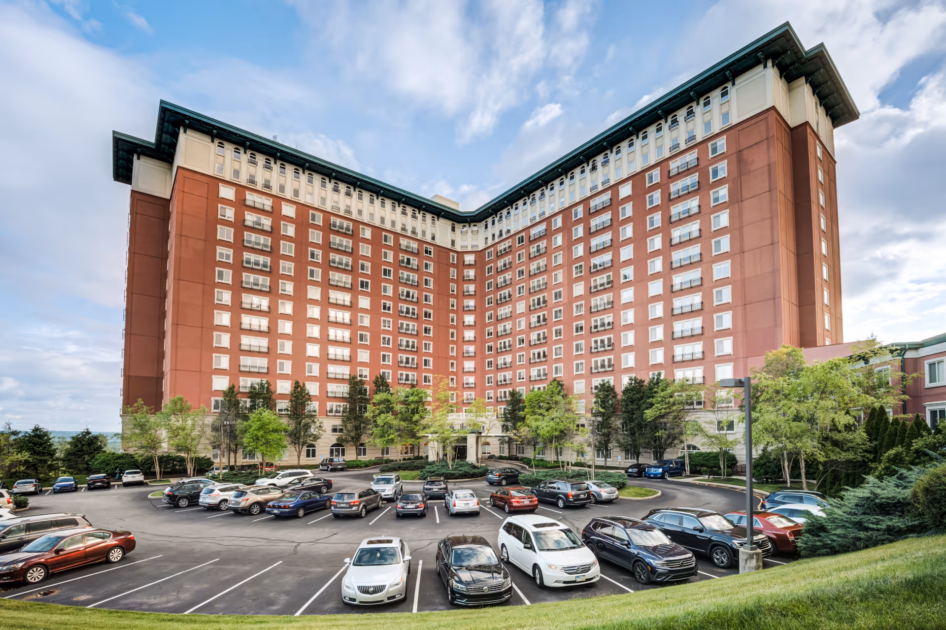 Large multi-story red-brick senior living building with many windows and a parking lot in front under a partly cloudy sky.