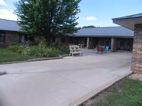 Outdoor courtyard area of Vernon Manor featuring a concrete patio with a bench decorated with sunflowers, several chairs and tables, a large tree, and a garden bed with plants. The building with a metal roof and brick walls surrounds the courtyard.