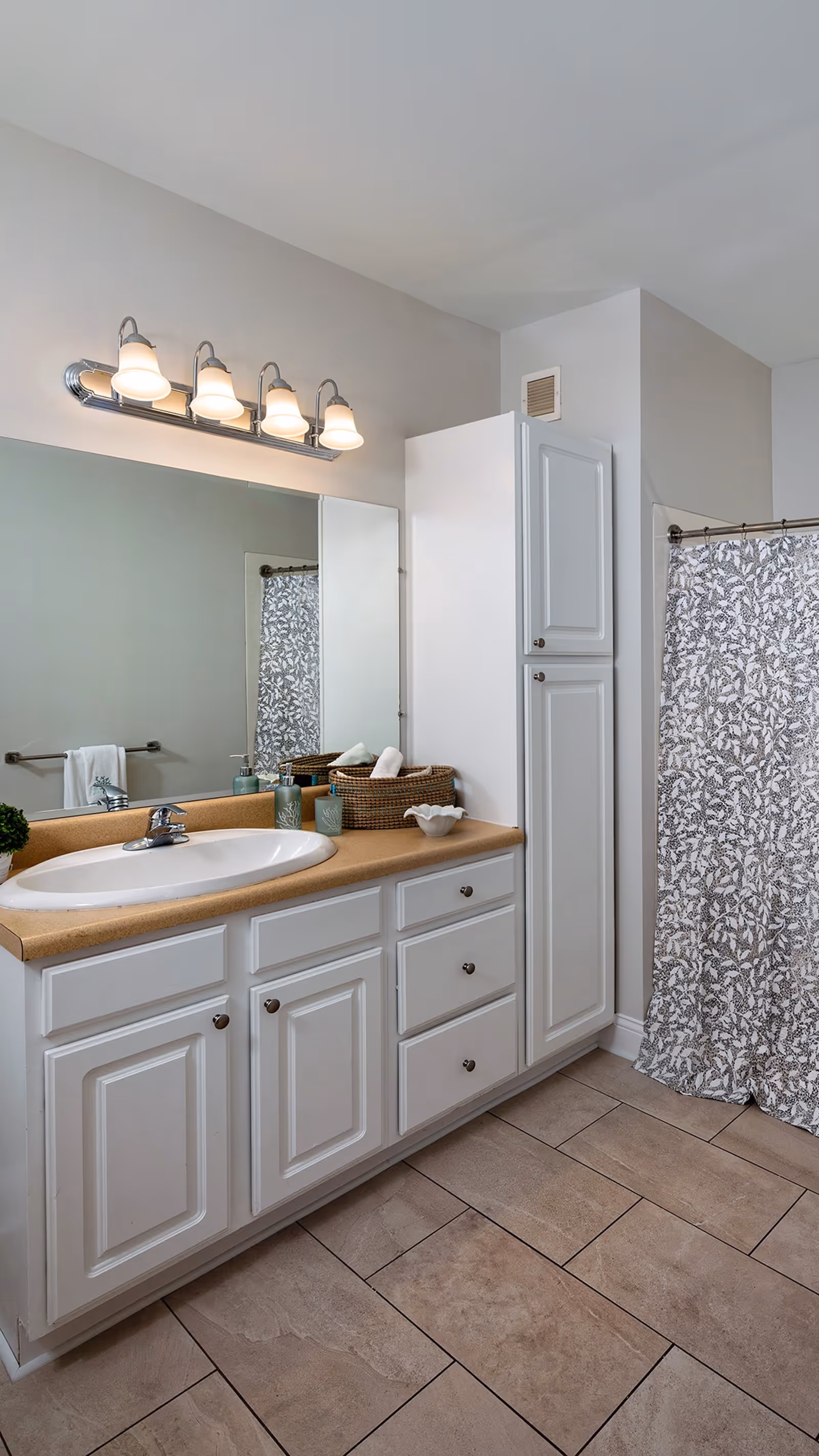 Well-lit bathroom with a white vanity and sink, large mirror, tall storage cabinet, and a patterned shower curtain.