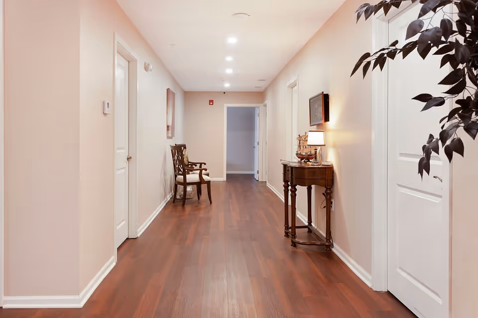 A long, well-lit interior hallway with polished wood floors, a chair and a small console table with a lamp.