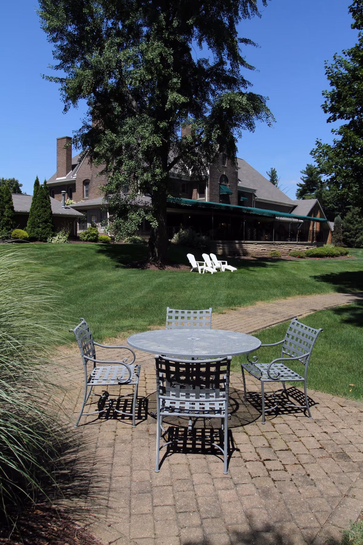 Outdoor patio area with a metal round table and four matching chairs on a brick-paved surface, surrounded by green grass and trees. In the background, there is a large brick building with a covered porch and two white Adirondack chairs on the lawn.