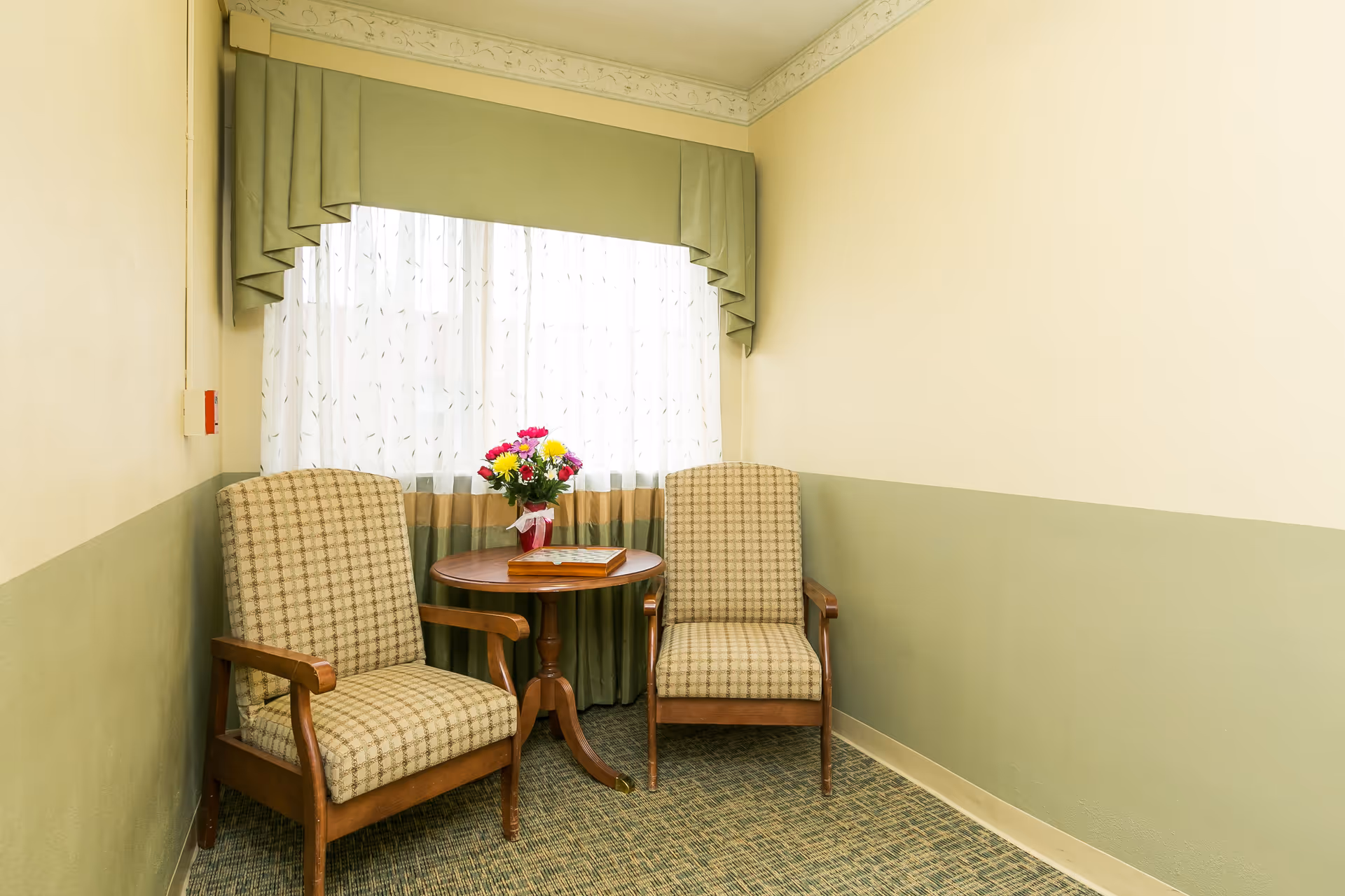 Two upholstered armchairs flank a small round table with a vase of flowers in front of a curtained window in a pale green interior.