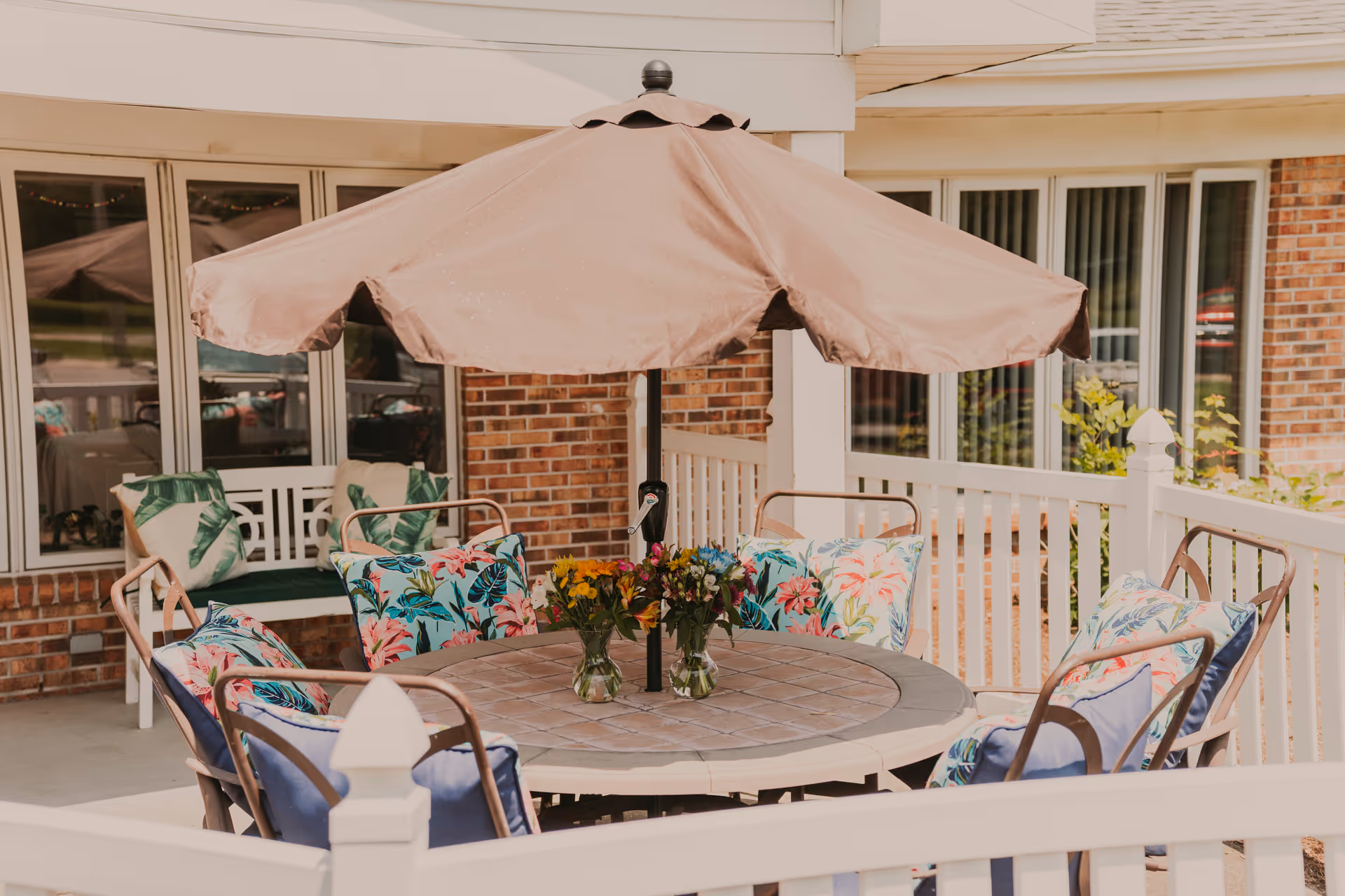 Outdoor patio area with a round table surrounded by chairs that have colorful floral cushions. A large beige umbrella is positioned in the center of the table, and two small vases with flowers are placed on the table. The patio is enclosed by a white railing and is adjacent to a brick building with windows.