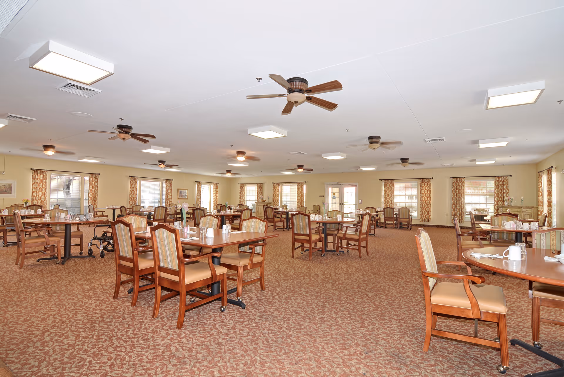 A spacious dining room with multiple wooden tables and chairs arranged neatly. The room has patterned carpet flooring, several ceiling fans, and large windows with patterned curtains allowing natural light to enter. Tables are set with cups, glasses, and napkins, and a walker is visible near one of the tables.
