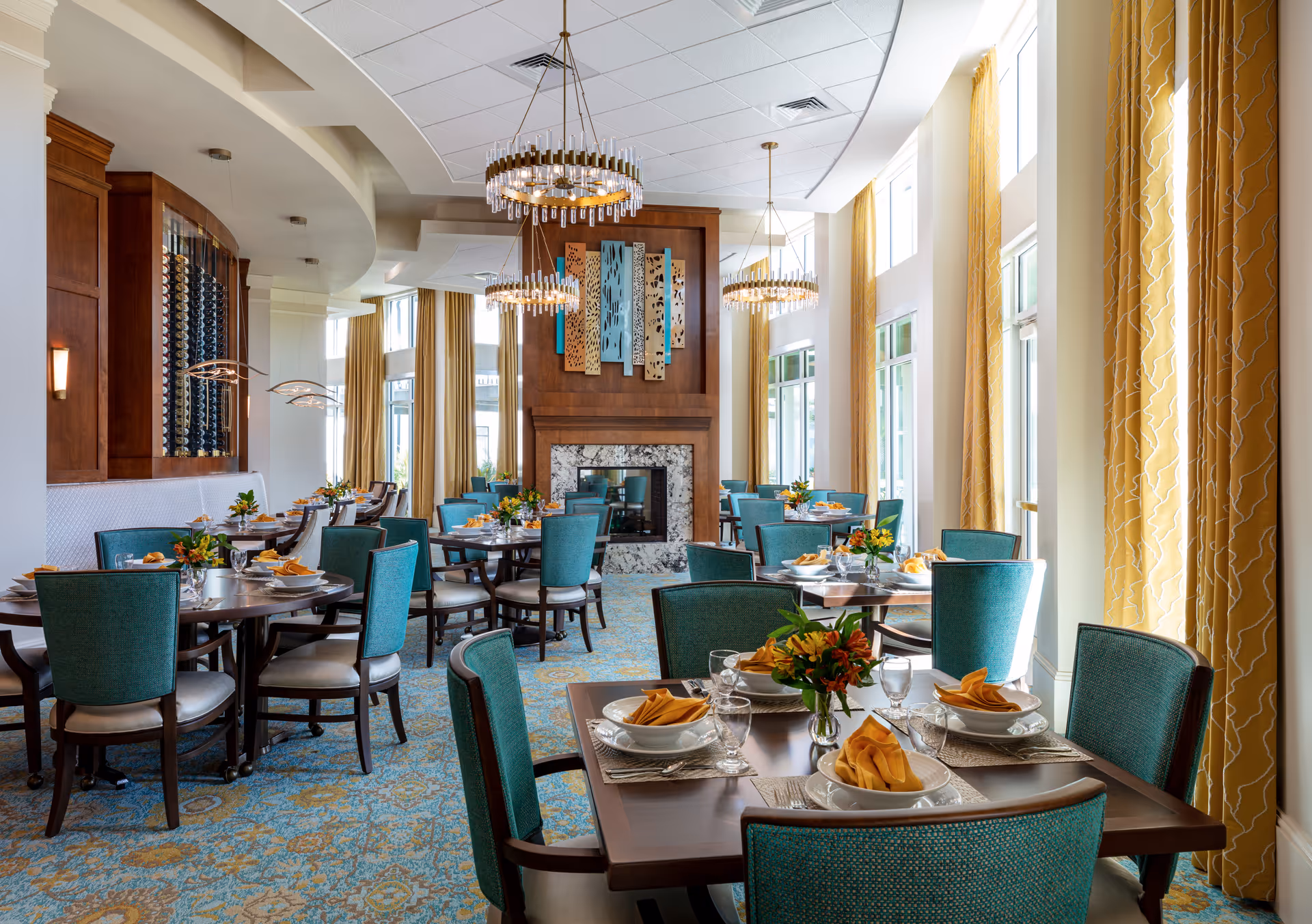 Bright, elegant dining room with teal chairs and round tables set with place settings, yellow curtains, chandeliers, and a central fireplace.