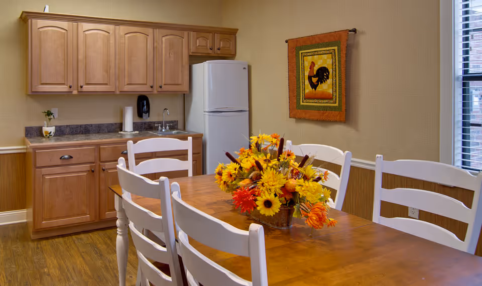 Small dining area with a wooden table and white chairs centered around a bright floral centerpiece, with a kitchenette, cabinets and refrigerator in the background.
