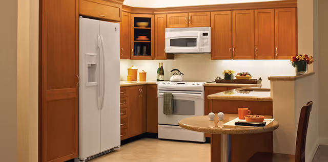 A modern kitchen with wooden cabinets, a white refrigerator, a white stove with a microwave above it, and a small round table with two chairs. The countertop has a few decorative items including a teapot, a bowl of fruit, and some containers.