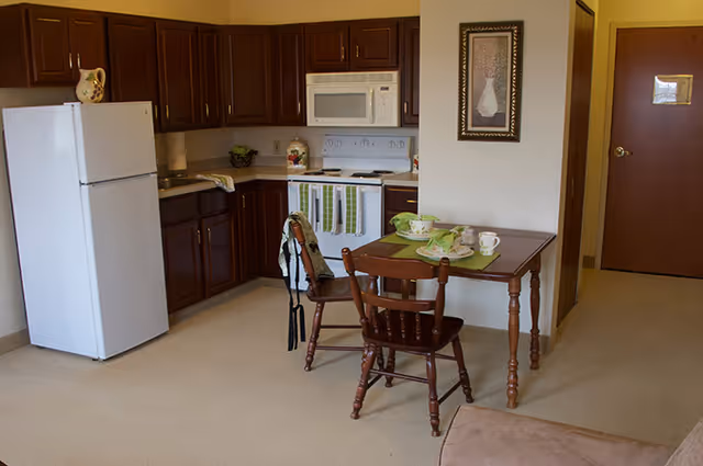 Small kitchenette with a white refrigerator, stove and microwave beside a wooden dining table set with two chairs in a senior living apartment.