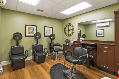Interior of a salon room with three black hair drying chairs against a green wall, a large wall clock, framed artwork, a wooden cabinet with a mirror, and a salon styling chair on a wooden floor.