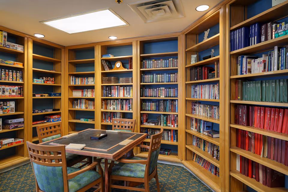 A cozy library room with wooden bookshelves filled with books and board games. In the center, there is a square table with a black playing surface and four wooden chairs with green patterned cushions. The room has a carpeted floor and recessed ceiling lights.