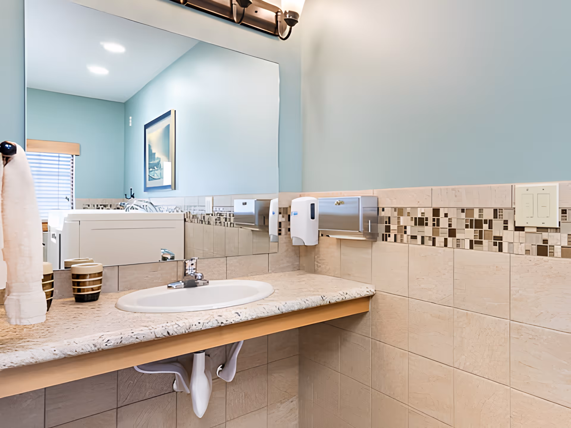 A clean bathroom sink area with a large mirror above a beige marble countertop. The walls are painted light blue with beige tiles and a decorative mosaic tile border. There is a towel hanging on the left side, a soap dispenser, and a stainless steel paper towel holder mounted on the wall.