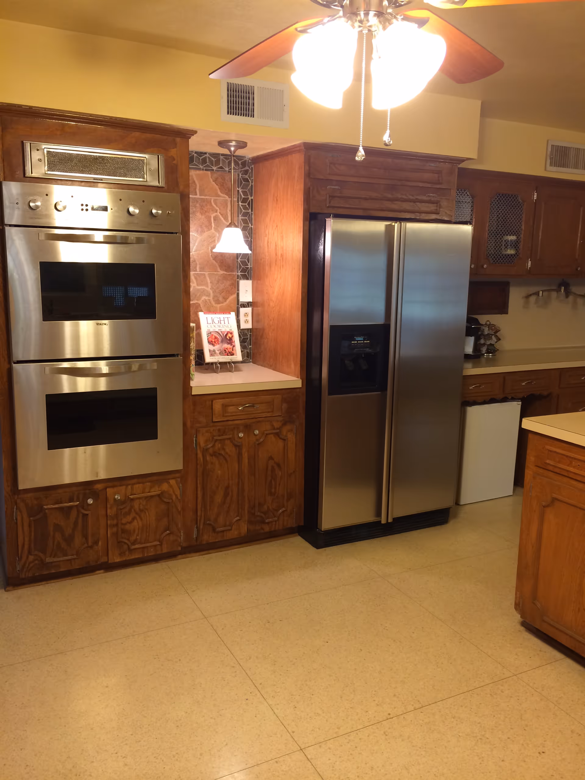 A kitchen with wooden cabinets, a double stainless steel oven, a stainless steel side-by-side refrigerator with a water and ice dispenser, a small lamp on the countertop, and a ceiling fan with lights above.