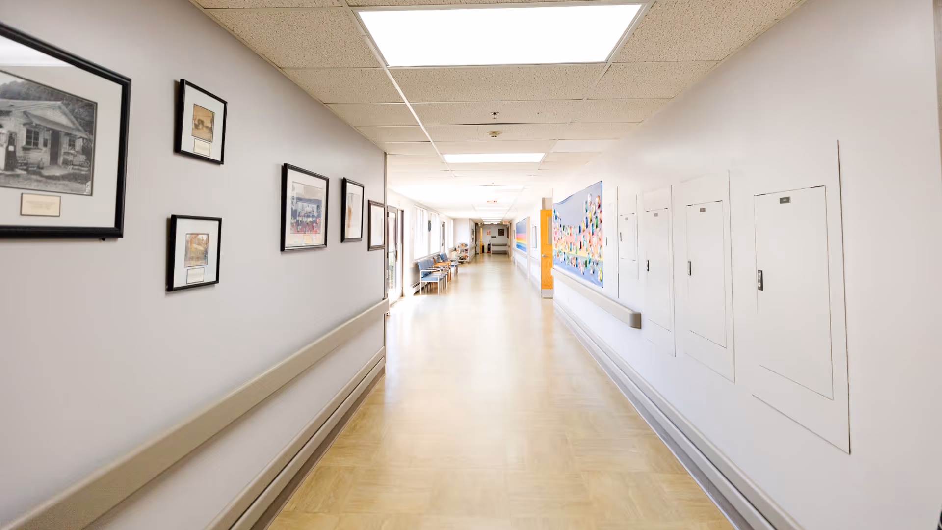 A long, well-lit hallway in a healthcare facility with framed pictures on the left wall and utility panels on the right wall. There are chairs and a bulletin board further down the corridor.