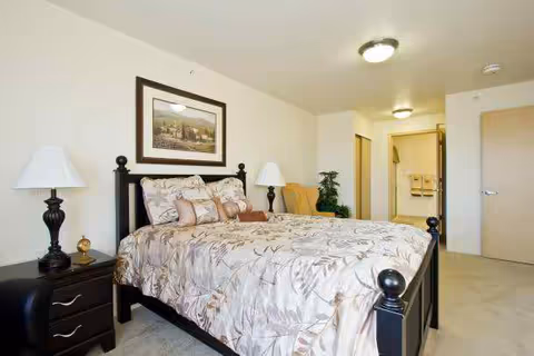 A neatly made bed with floral patterned bedding in a bedroom. The bed has a black frame and is flanked by two dark nightstands, each with a lamp. A framed picture hangs on the wall above the bed. In the background, there is an open doorway leading to a bathroom with towels hanging on the wall and a plant near the entrance.