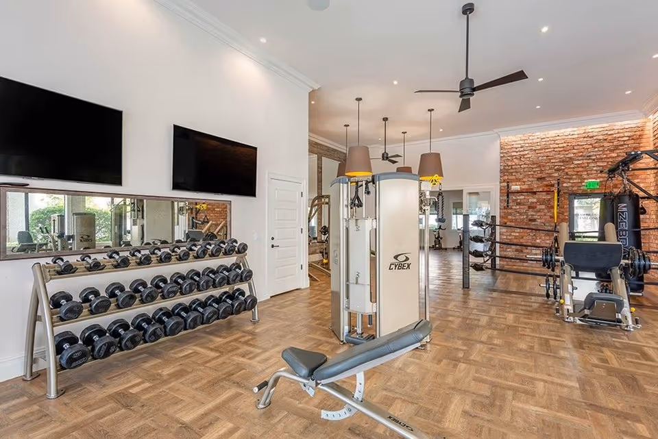 Modern fitness room with a rack of dumbbells, weight machines, TVs, and an exposed brick wall.