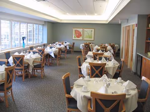 A dining room with multiple round tables covered with white tablecloths, each set with cups, saucers, napkins, and silverware. Wooden chairs with cushioned seats surround the tables. Large windows on the left side let in natural light, and two framed floral artworks hang on the far wall.