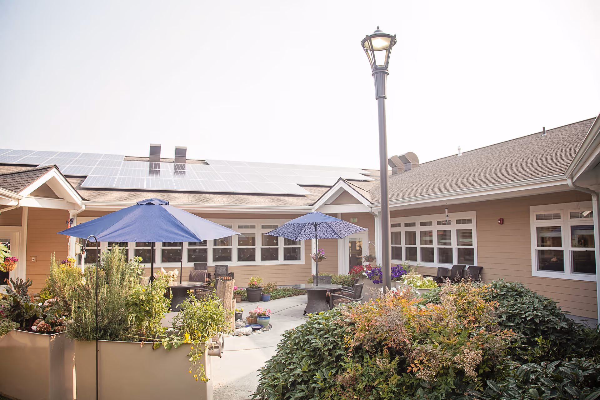 Outdoor courtyard area of a senior living facility with beige buildings, solar panels on the roof, two blue umbrellas shading tables and chairs, various potted plants and flowers, and a tall street lamp in the center.