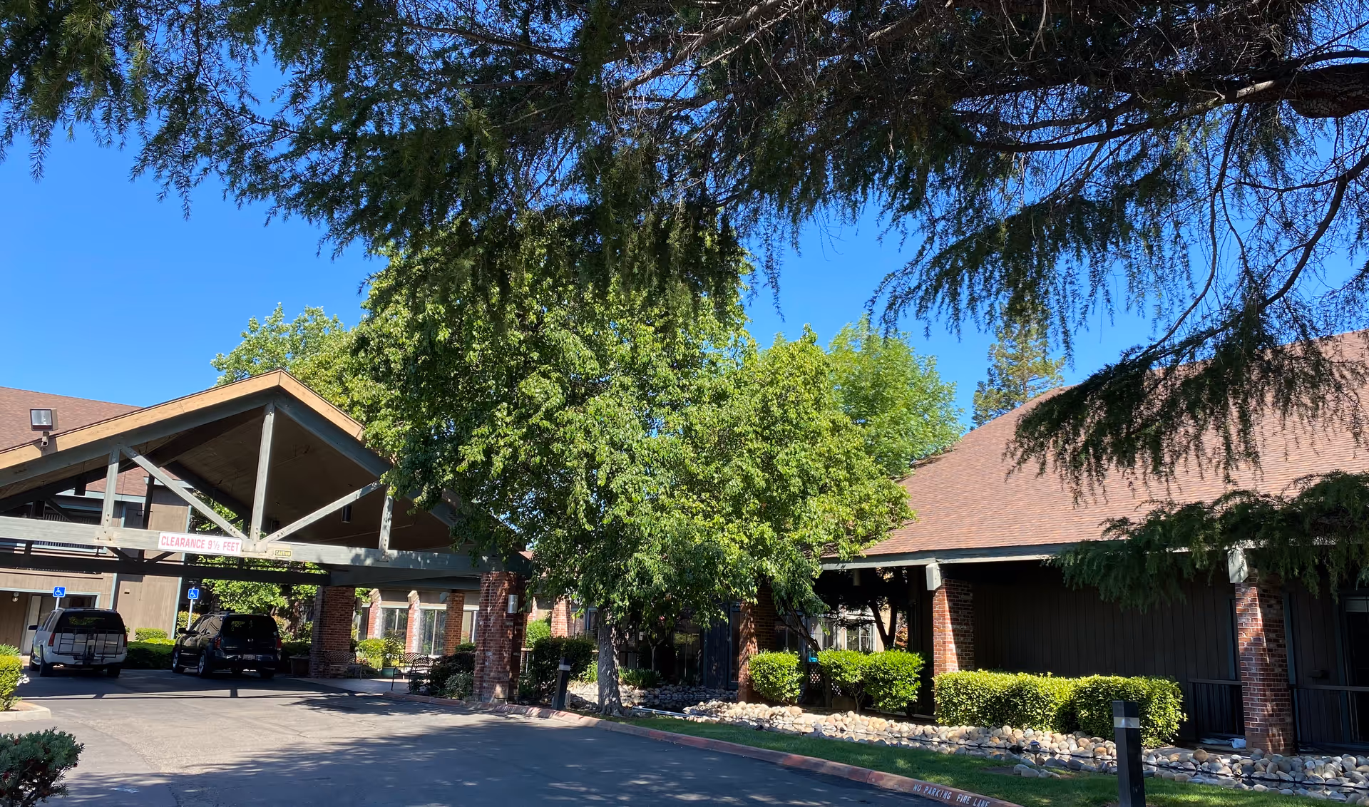 Exterior view of The Crest at Citrus Heights facility showing a covered driveway entrance with clearance sign, surrounded by trees and bushes under a clear blue sky.