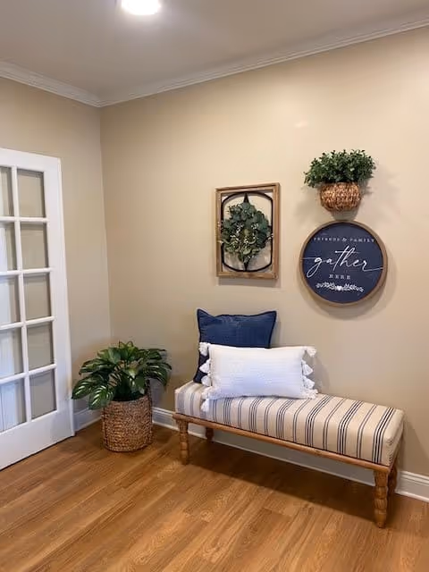A cozy corner with a striped cushioned bench adorned with two pillows, one navy blue and one white. Above the bench, there are three wall decorations: a framed green wreath, a round sign that says 'gather', and a small hanging basket with greenery. To the left, there is a potted plant in a woven basket on a wooden floor, next to a white-framed glass door.