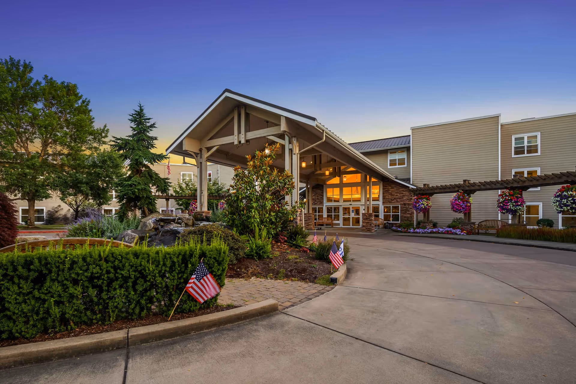 Exterior view of Crista Shores senior living facility at dusk, showing the main entrance with a covered driveway, landscaped garden with bushes, flowers, small American flags, and trees surrounding the building.