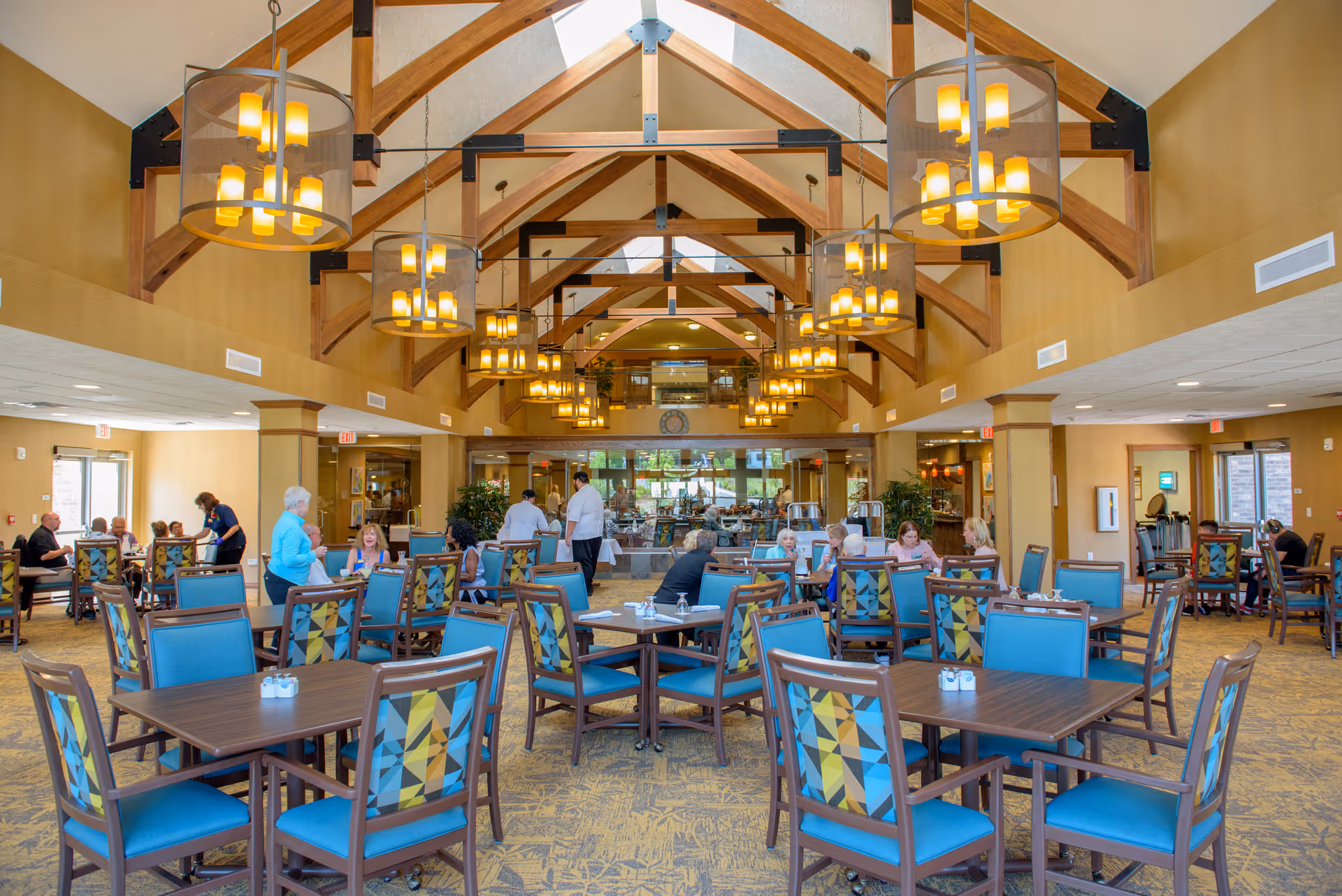 Spacious dining room in a retirement resort with wooden tables and chairs featuring blue and patterned upholstery. Several people are seated and interacting, with staff attending to guests. The room has high vaulted ceilings with exposed wooden beams and multiple hanging light fixtures, creating a warm and inviting atmosphere.
