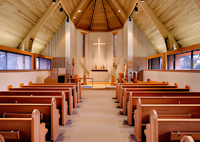 Interior view of a chapel with wooden pews arranged in rows facing an altar with a glowing cross on the wall behind it. The ceiling is vaulted with wooden beams and there are narrow windows along the sides.