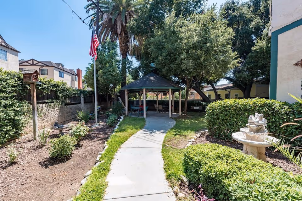 Paved walkway through a landscaped courtyard with a gazebo, American flag, fountain and surrounding trees and shrubs.