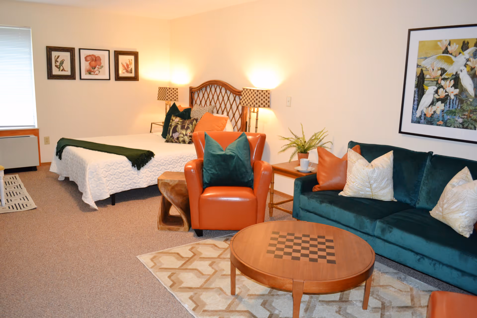 Studio apartment interior with a bed in the background, green velvet sofa, orange armchair, and wooden coffee table.