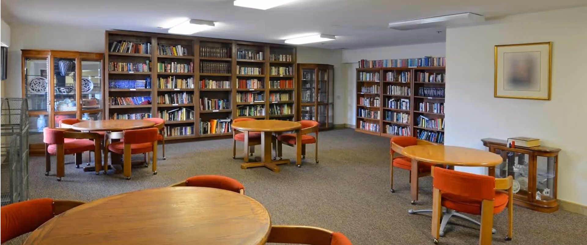 A quiet library room with multiple wooden round tables surrounded by red cushioned chairs. The walls are lined with tall bookshelves filled with books, and there are glass display cabinets on the left and right sides. The room is carpeted and well-lit with ceiling lights.