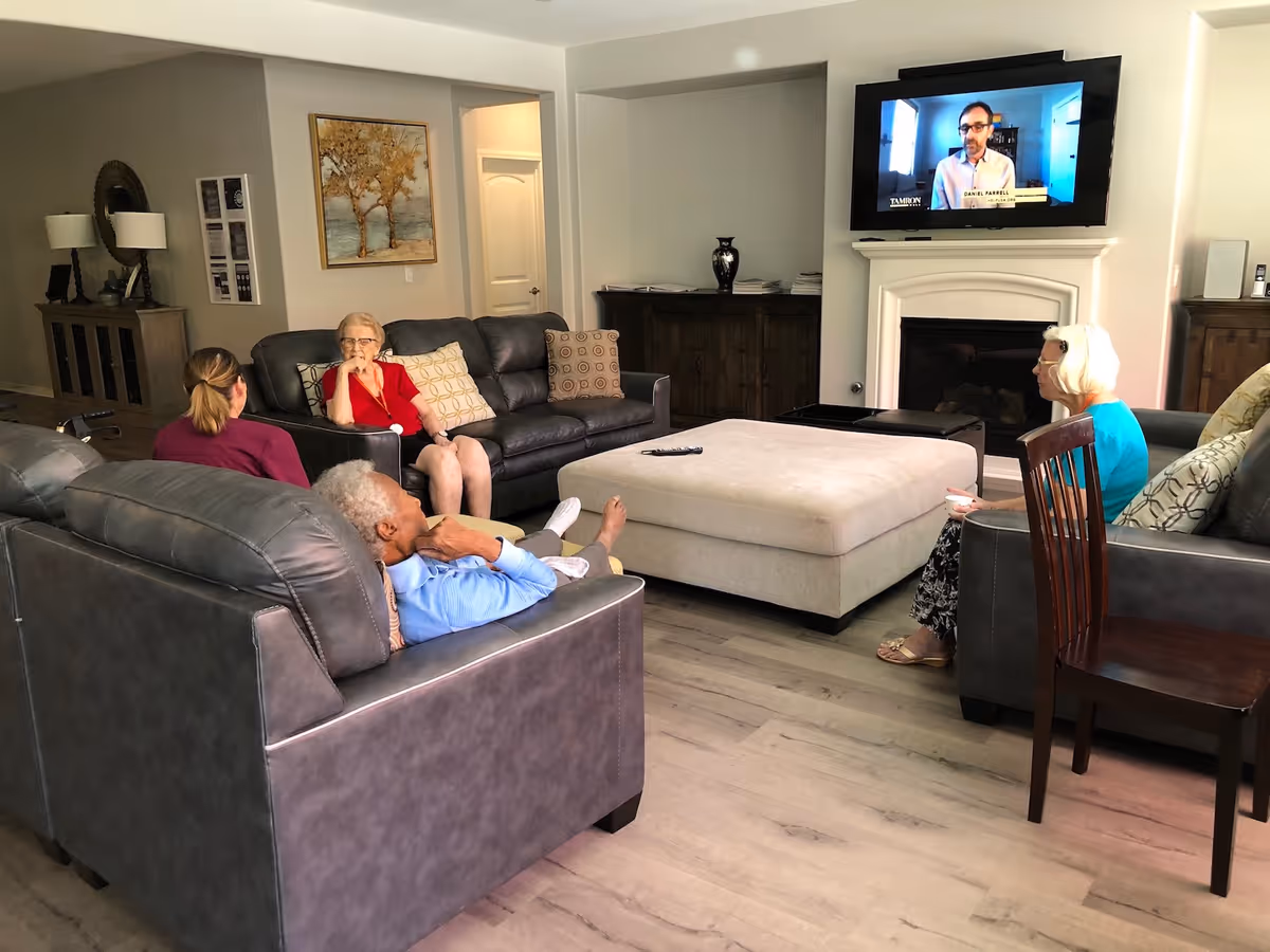 A living room with four elderly people seated on sofas and chairs, watching a television mounted above a fireplace. The room has wooden flooring, a large beige ottoman in the center, and decorative items including a painting, lamps, and vases.