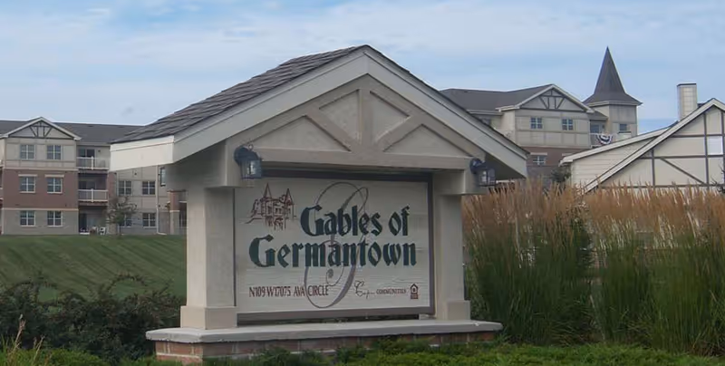 Outdoor view of the entrance sign for Gables of Germantown with a large building complex in the background under a partly cloudy sky.